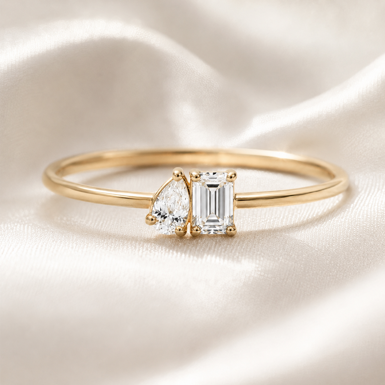 A gold ring featuring a pear-cut diamond and an emerald-cut diamond, displayed on a soft, draped white satin fabric.