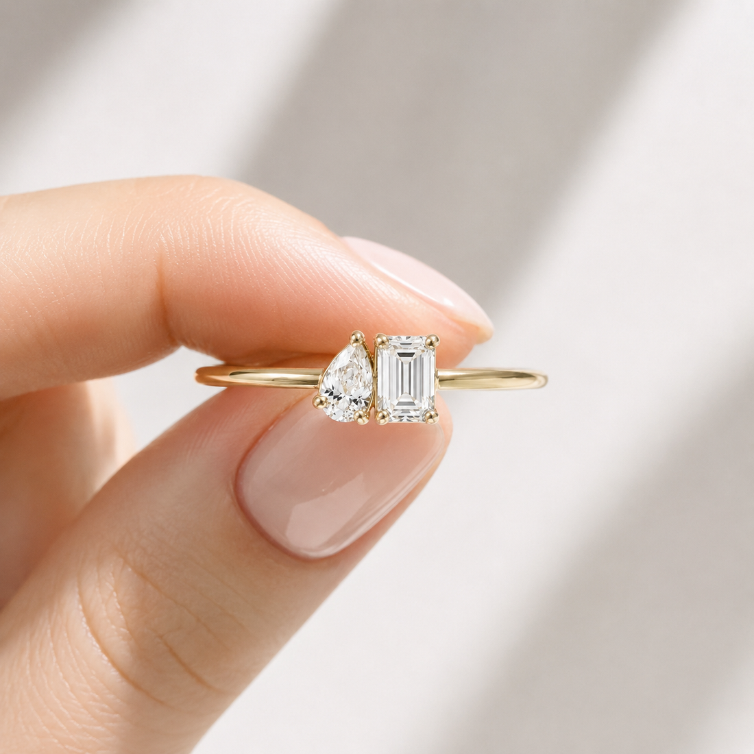 A gold ring with a pear-cut diamond and an emerald-cut diamond held between fingers against a soft, light gray background.