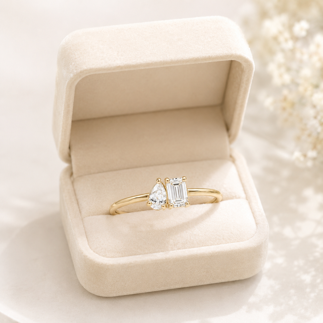 A gold ring with a pear-cut clear gemstone and an emerald-cut clear gemstone, displayed inside an open cream-colored velvet ring box.