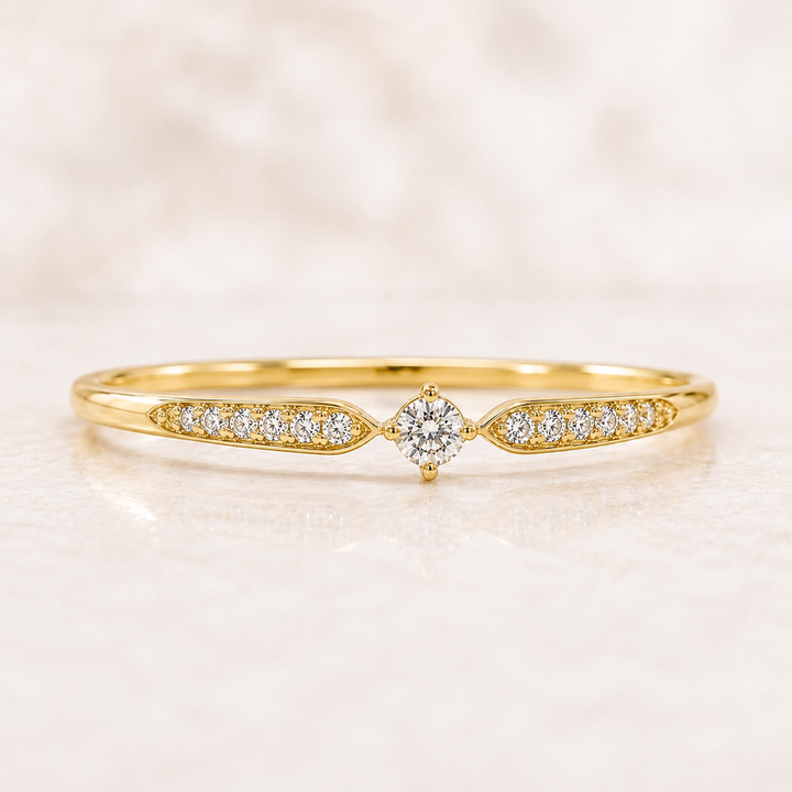 A delicate yellow gold ring featuring a central round-cut diamond flanked by pavé diamond sections on the band, set against a soft, light marble background.