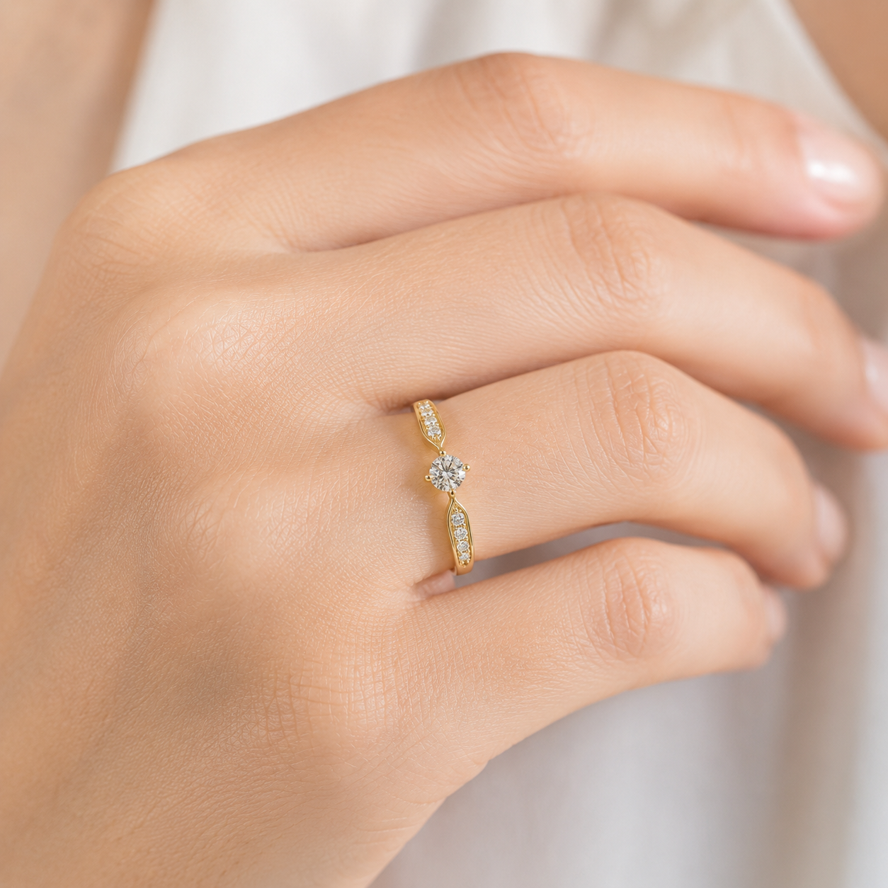 A close-up of a hand wearing a delicate yellow gold ring with a central clear gemstone and small side stones, set against a soft white background.