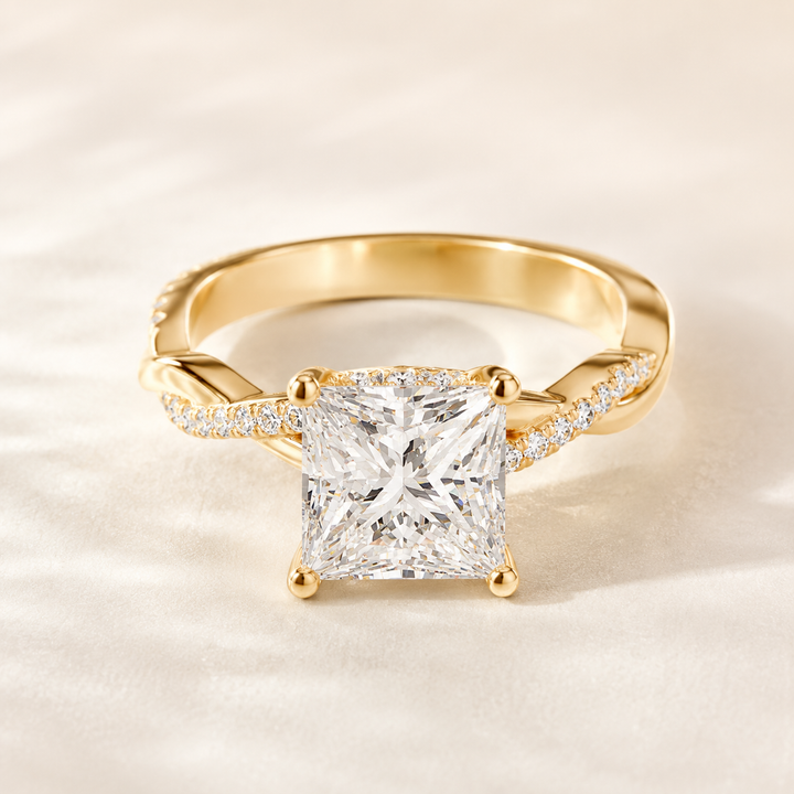 A yellow gold ring featuring a large princess-cut diamond center stone and pavé diamonds along the twisted band, set against a soft, light beige fabric background.