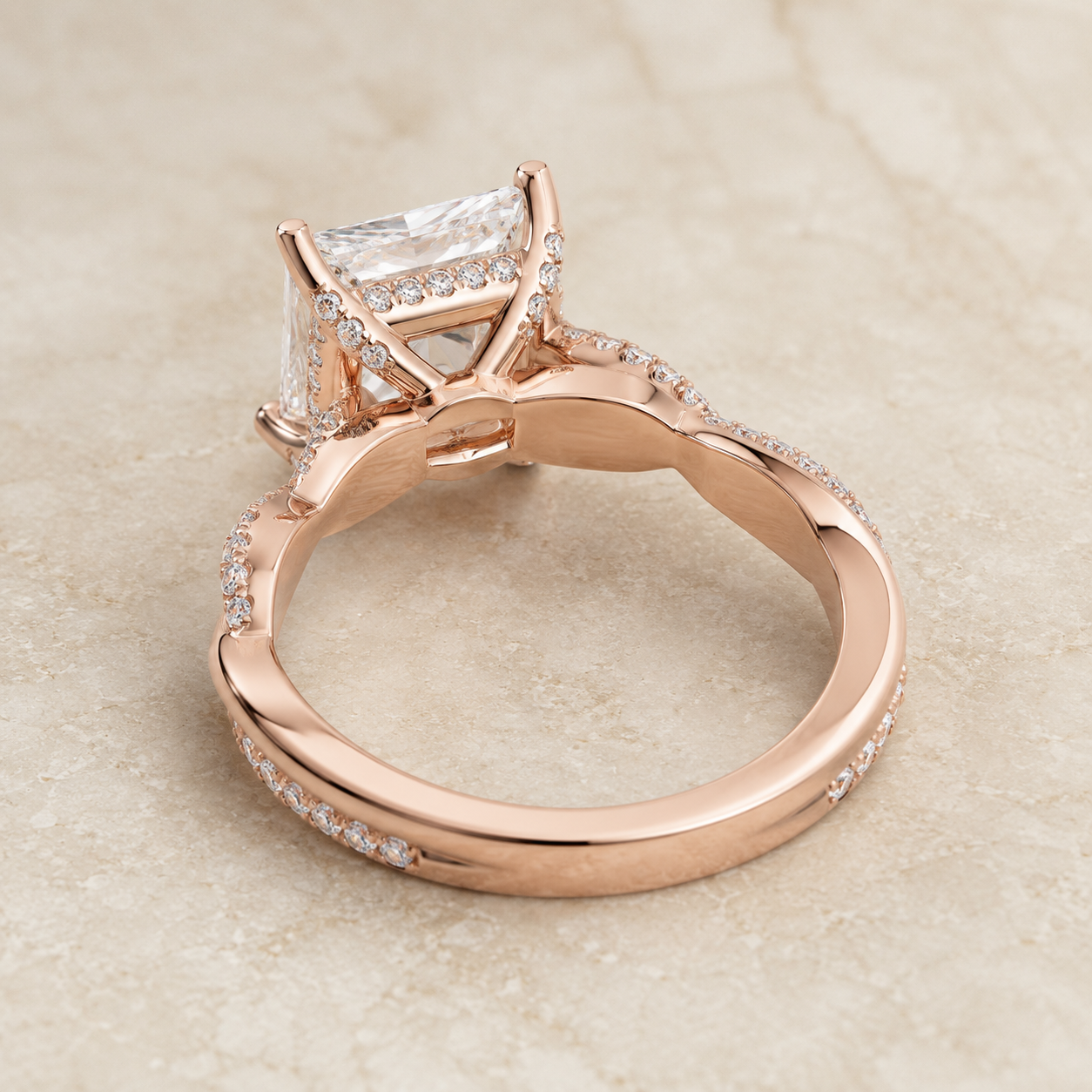 Rose gold ring with a rectangular clear gemstone and pavé accents, photographed on a light beige stone surface.
