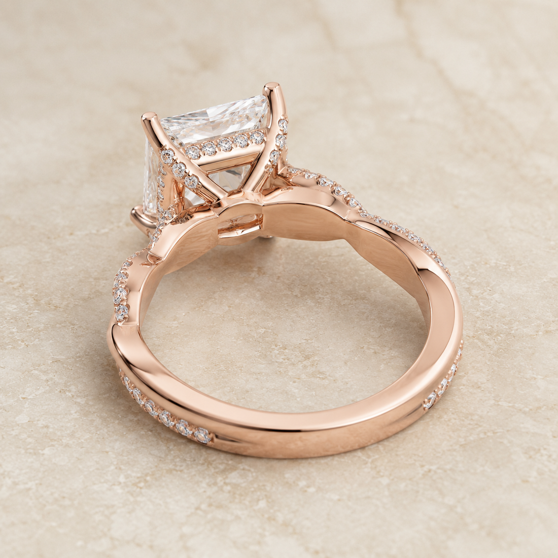 Rose gold ring with a rectangular clear gemstone and pavé accents, photographed on a light beige stone surface.