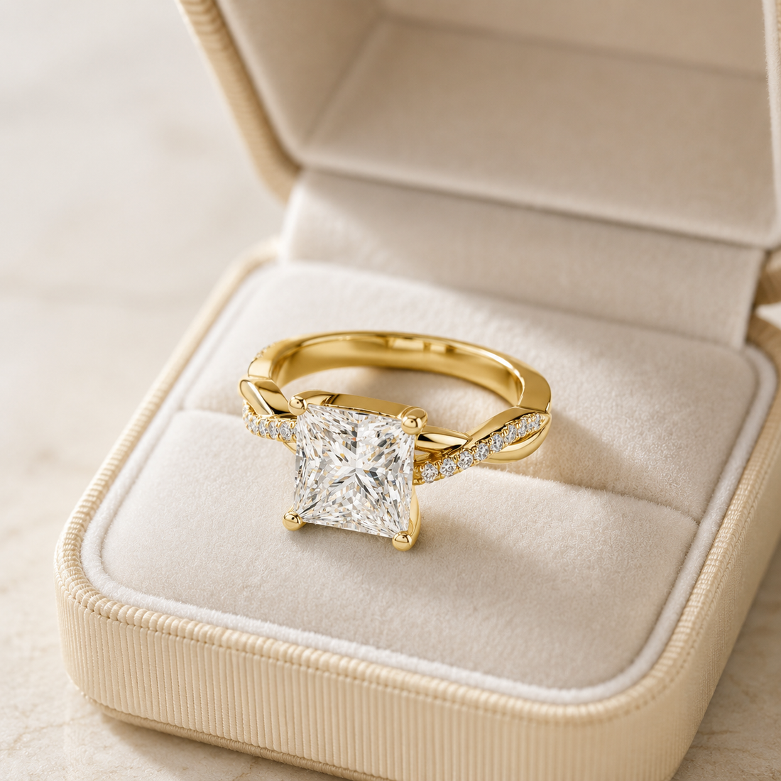 A yellow gold ring with a princess-cut diamond solitaire and small pavé diamonds on the band, displayed inside an open cream-colored jewelry box.