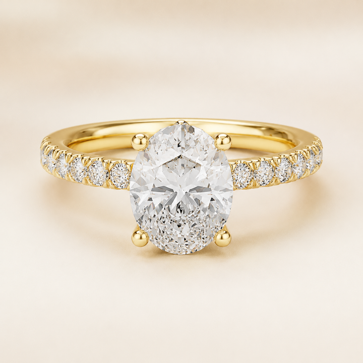 A yellow gold engagement ring featuring a large oval-cut center stone in a four-prong setting, with a pavé band set with small round diamonds, shown on a soft beige background.