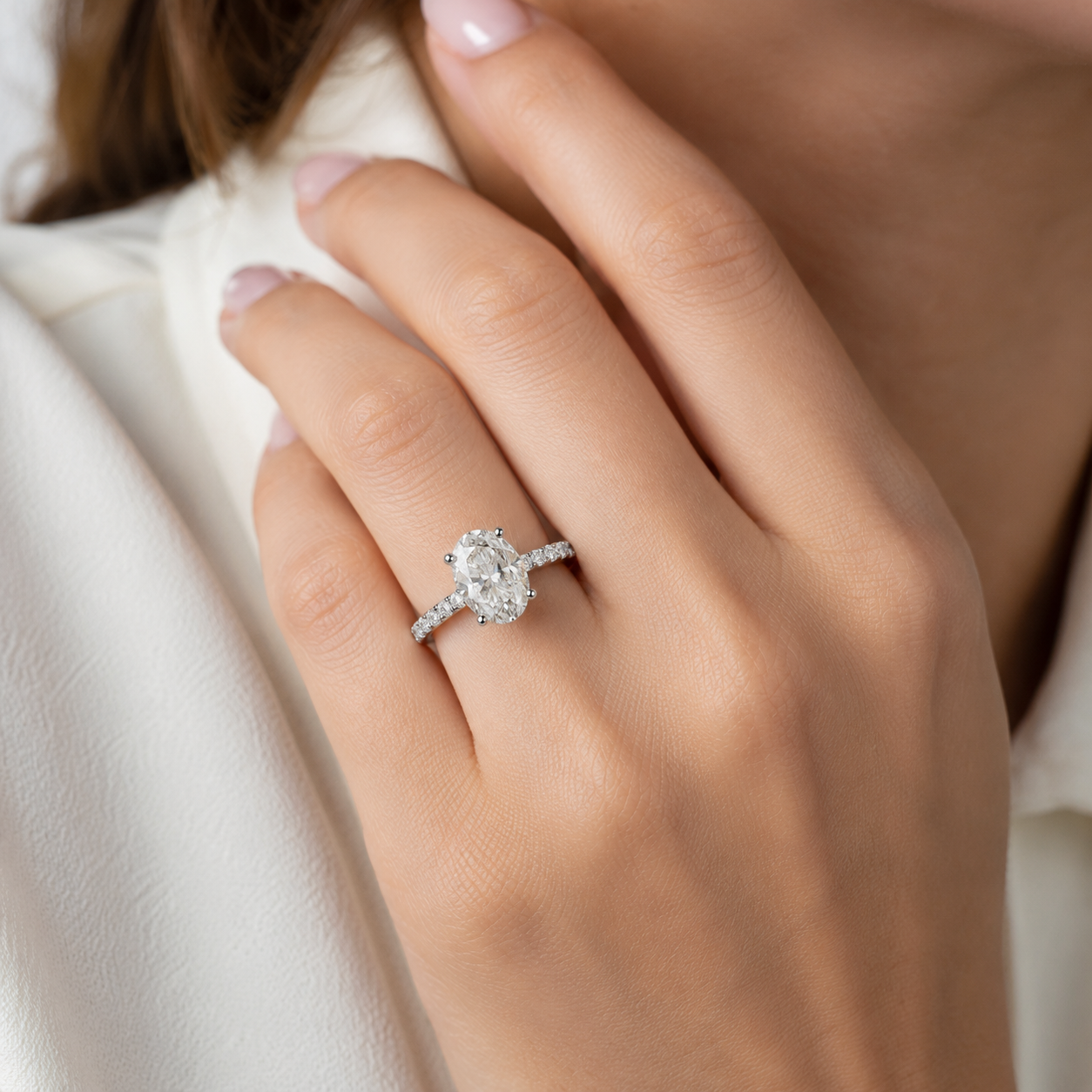 A close-up of a woman's hand wearing a silver-toned diamond engagement ring with an oval center stone and pavé band, set against a soft cream-colored blouse.