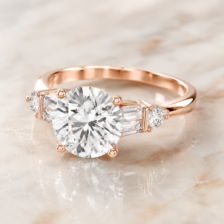 A rose gold engagement ring featuring a large round-cut center diamond flanked by two baguette-cut side diamonds and small round accent diamonds, set on a polished band, resting on a light marble surface.
