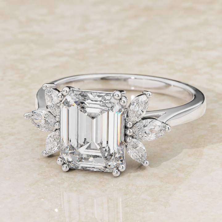 An emerald-cut diamond ring in a silver-colored metal setting, featuring marquise-cut side diamonds, photographed on a light beige textured surface.