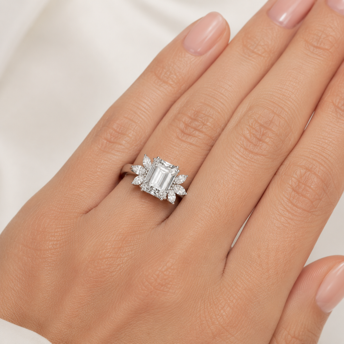 A close-up of a hand wearing a silver ring with an emerald-cut center stone and smaller surrounding stones, photographed against a soft light background.