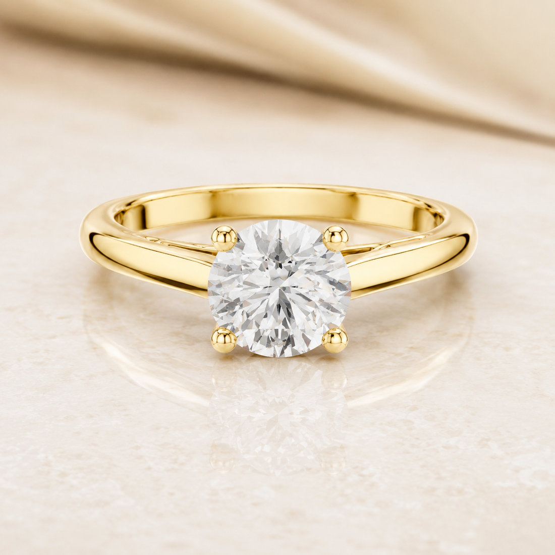 A yellow gold solitaire engagement ring with a round brilliant-cut diamond set in a four-prong setting, resting on a light beige marble surface with a draped satin fabric in the background.