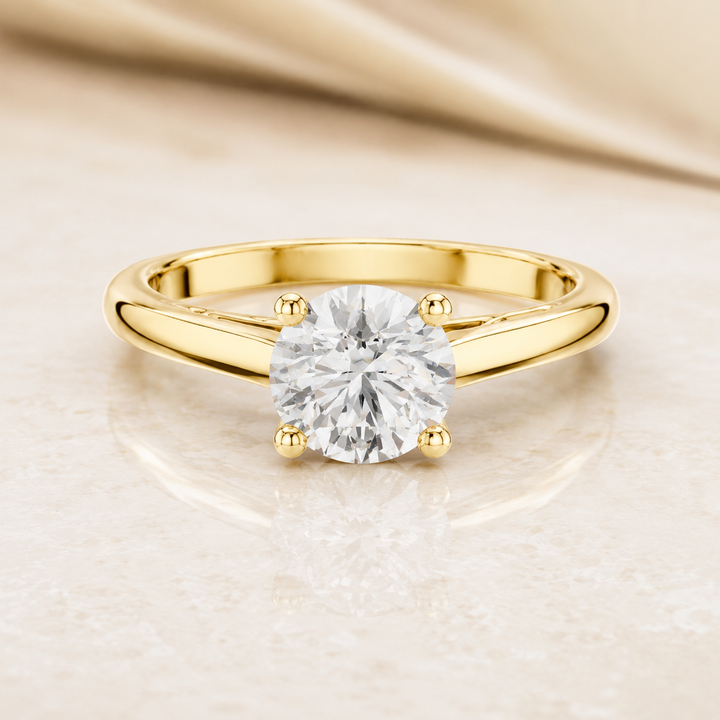 A yellow gold solitaire engagement ring with a round brilliant-cut diamond set in a four-prong setting, resting on a light beige marble surface with a draped satin fabric in the background.