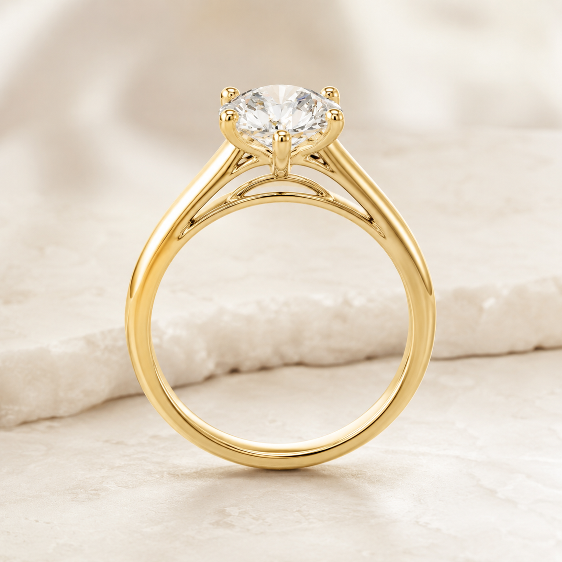 A yellow gold solitaire engagement ring with a round brilliant-cut diamond, displayed on a soft, textured white stone surface.