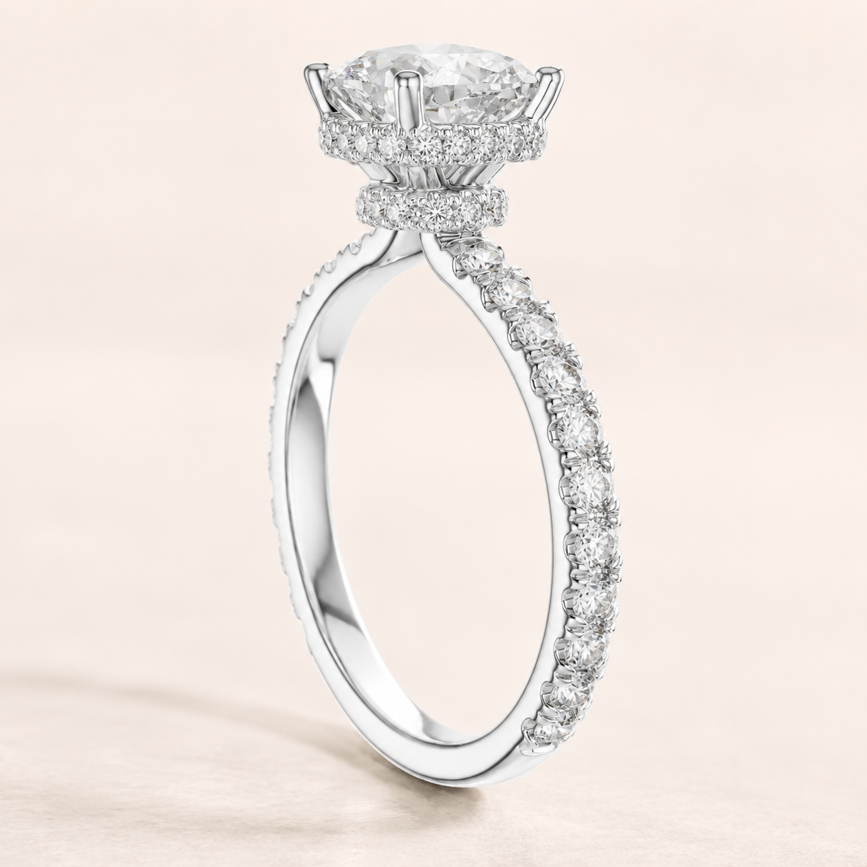 A radiant diamond ring in a polished silver-tone setting, featuring a large round center stone and a halo of smaller diamonds, with a pavé-set band, shown on a soft beige background.
