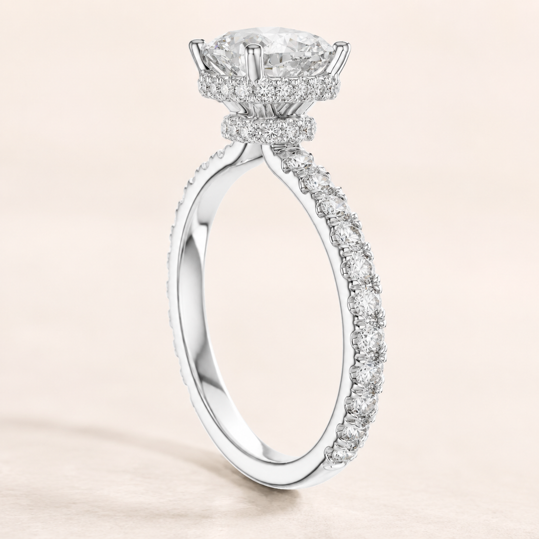 A radiant diamond ring in a polished silver-tone setting, featuring a large round center stone and a halo of smaller diamonds, with a pavé-set band, shown on a soft beige background.
