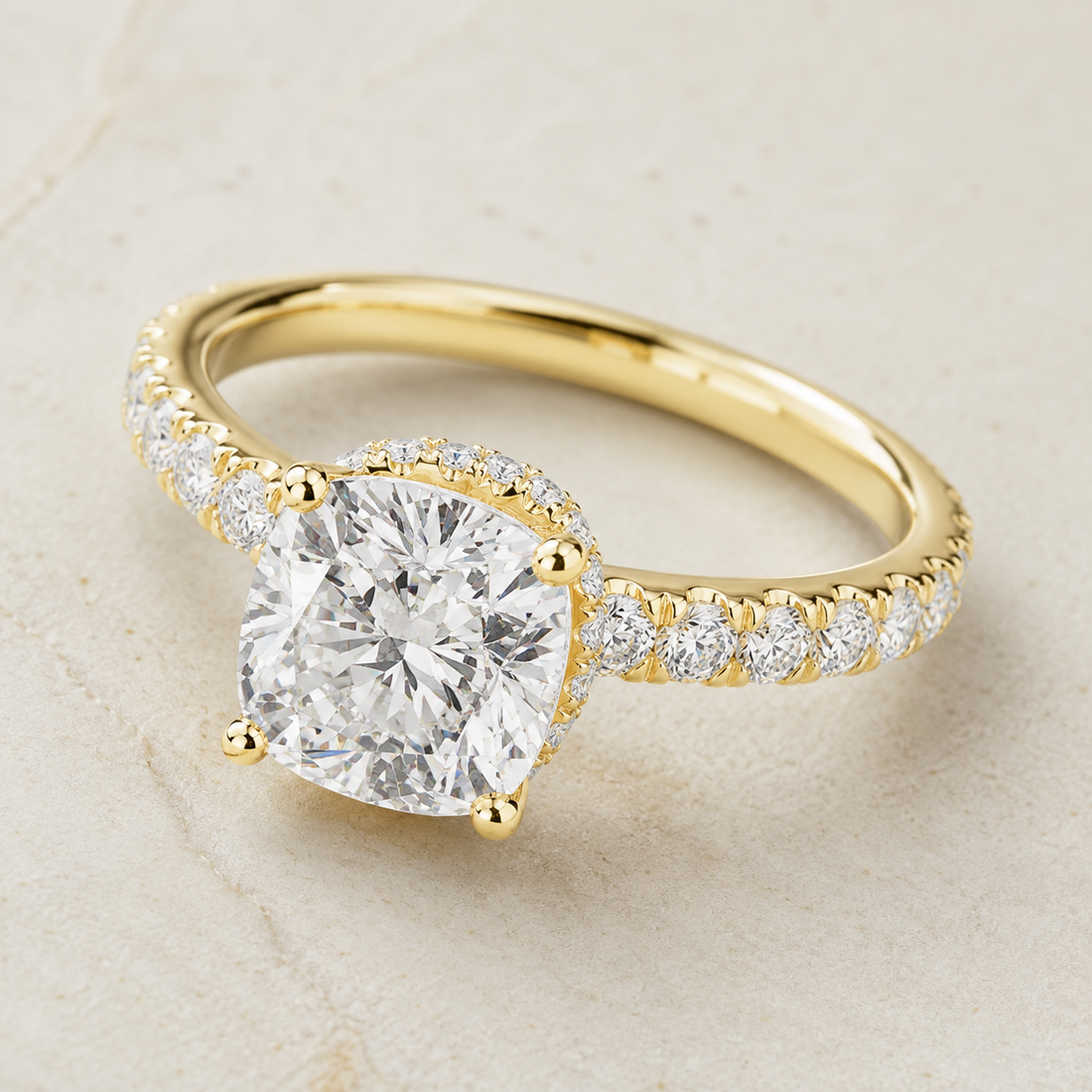A yellow gold engagement ring featuring a large cushion-cut diamond center stone and a pavé-set diamond band, shown on a light beige textured background.