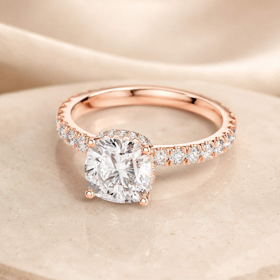 Rose gold engagement ring with a cushion-cut diamond center stone and pavé diamonds on the band, displayed on a beige stone surface.