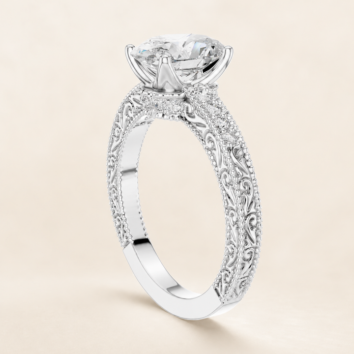 An ornate silver-toned diamond engagement ring with a large round-cut center stone and detailed filigree engraving on the shank, shown on a warm beige background.