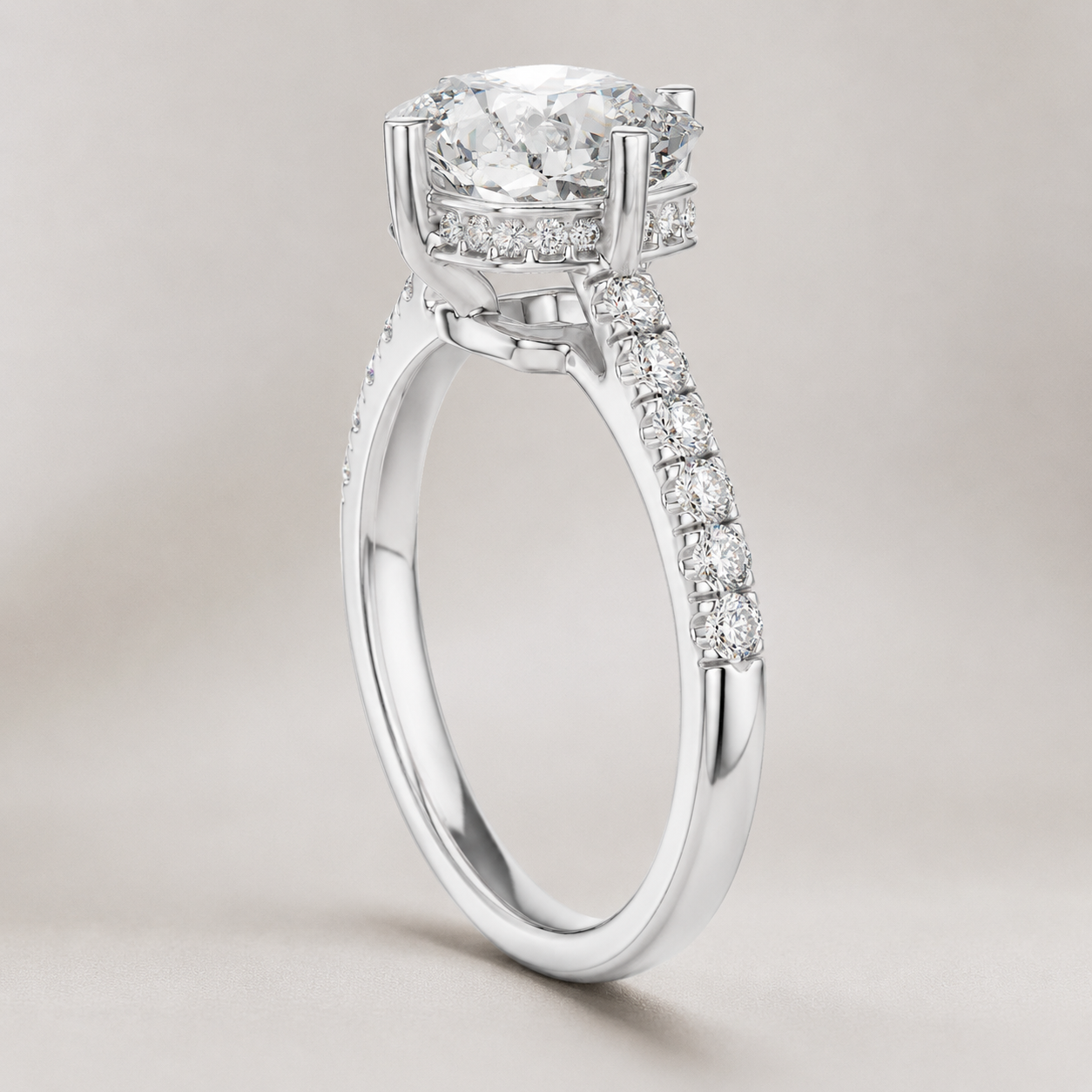 A silver-toned diamond ring with a round-cut center stone, a halo of smaller diamonds, and a pavé-set shank, shown on a neutral light gray background.