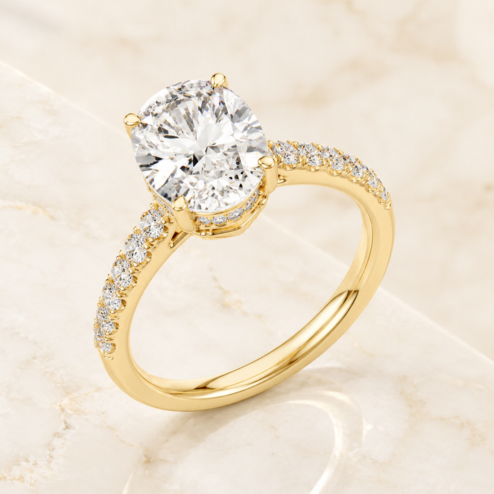 A yellow gold solitaire engagement ring with a round brilliant-cut center stone and a pavé-set diamond band, displayed on a light beige marble surface.