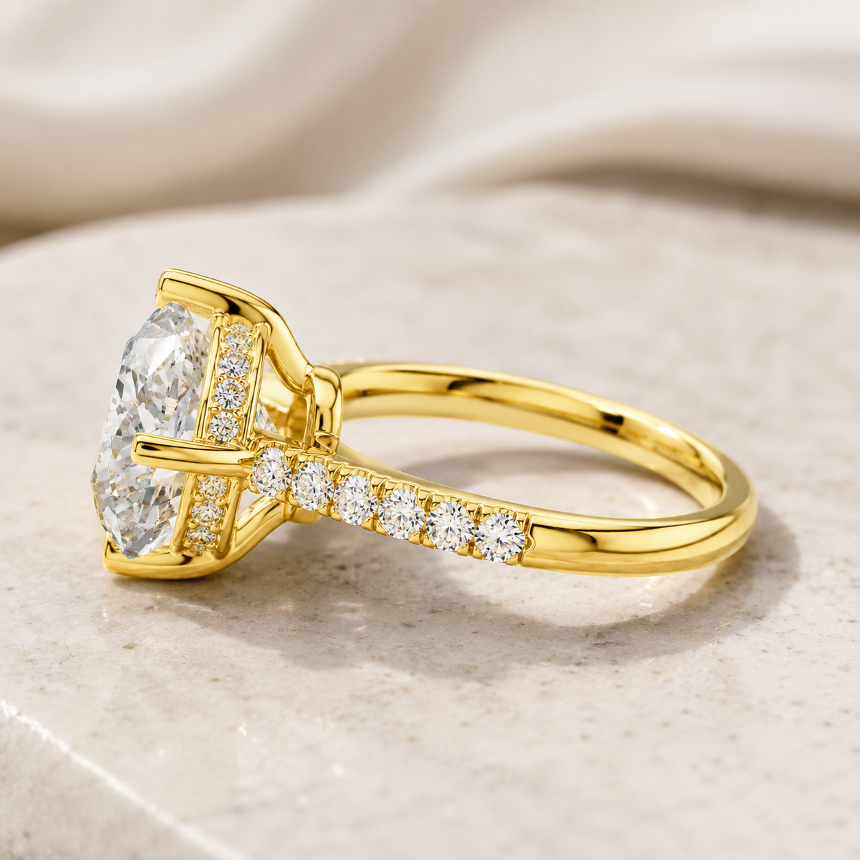 A yellow gold ring with a large oval-cut center stone and small pavé-set round stones along the shank, photographed on a soft beige fabric and stone surface.