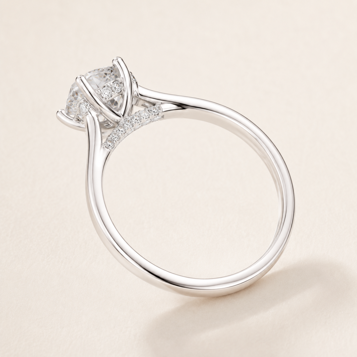 A silver-toned solitaire engagement ring with a round brilliant-cut center stone and small pavé accents on the shank, shown on a light beige background.