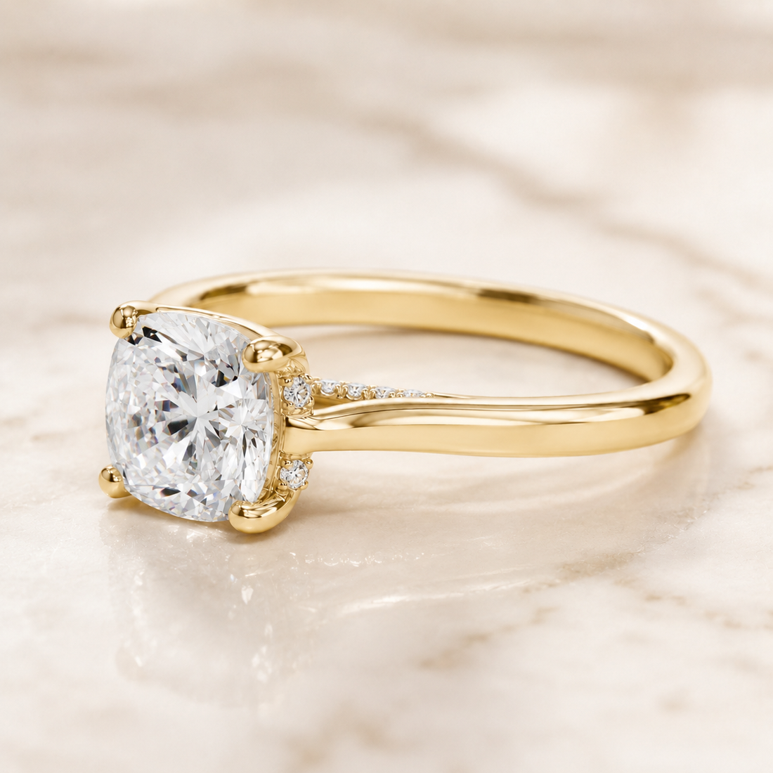 A yellow gold solitaire engagement ring with a cushion-cut diamond center stone and small accent diamonds on the band, resting on a light marble surface.