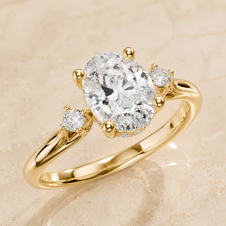 A yellow gold three-stone engagement ring with a large oval-cut center diamond and two smaller round diamonds on the shoulders, set against a light beige marble background.