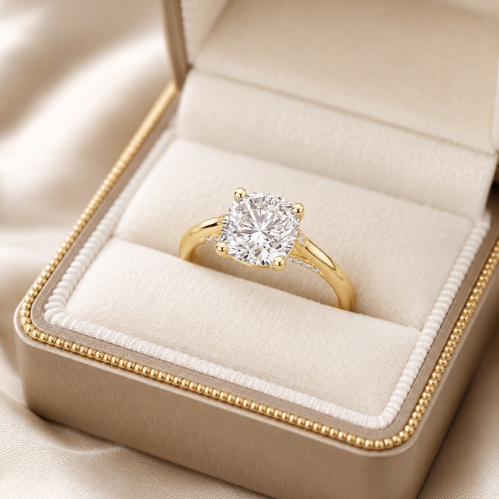 A yellow gold solitaire engagement ring with a round brilliant-cut diamond, displayed in an open champagne-colored velvet ring box with a gold beaded trim.