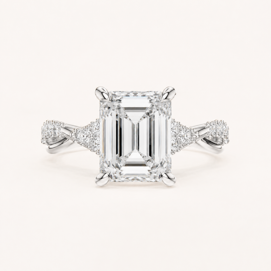 An emerald-cut diamond engagement ring in a silver-toned setting with pavé accents on the shoulders, centered on a light cream background.