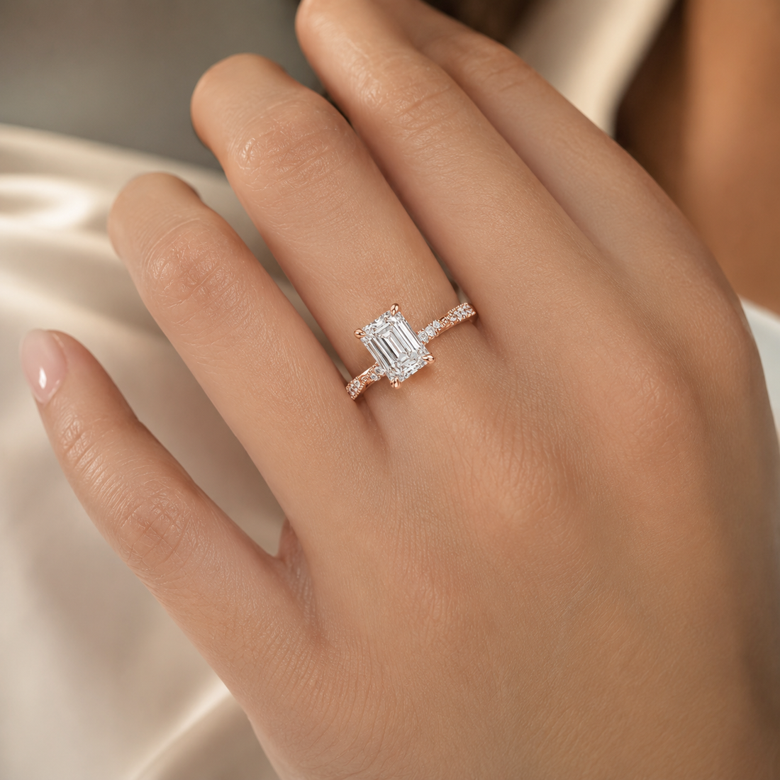 A close-up of a hand wearing a silver engagement ring with an emerald-cut center stone and a pavé band, set against a soft cream satin background.