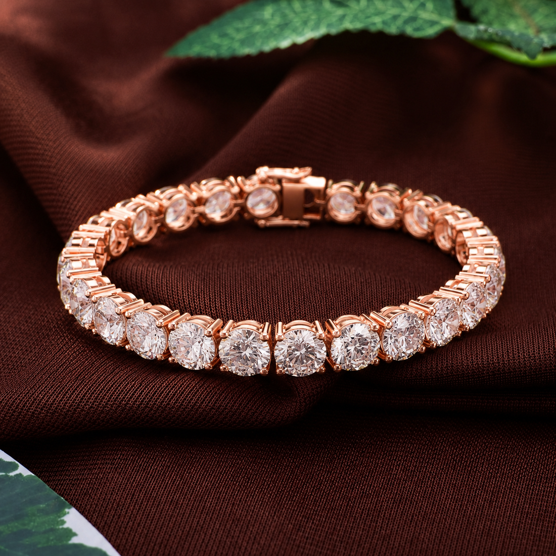 A rose gold tennis bracelet with round clear gemstones, displayed on a draped dark brown fabric background with green leaves in the corner.