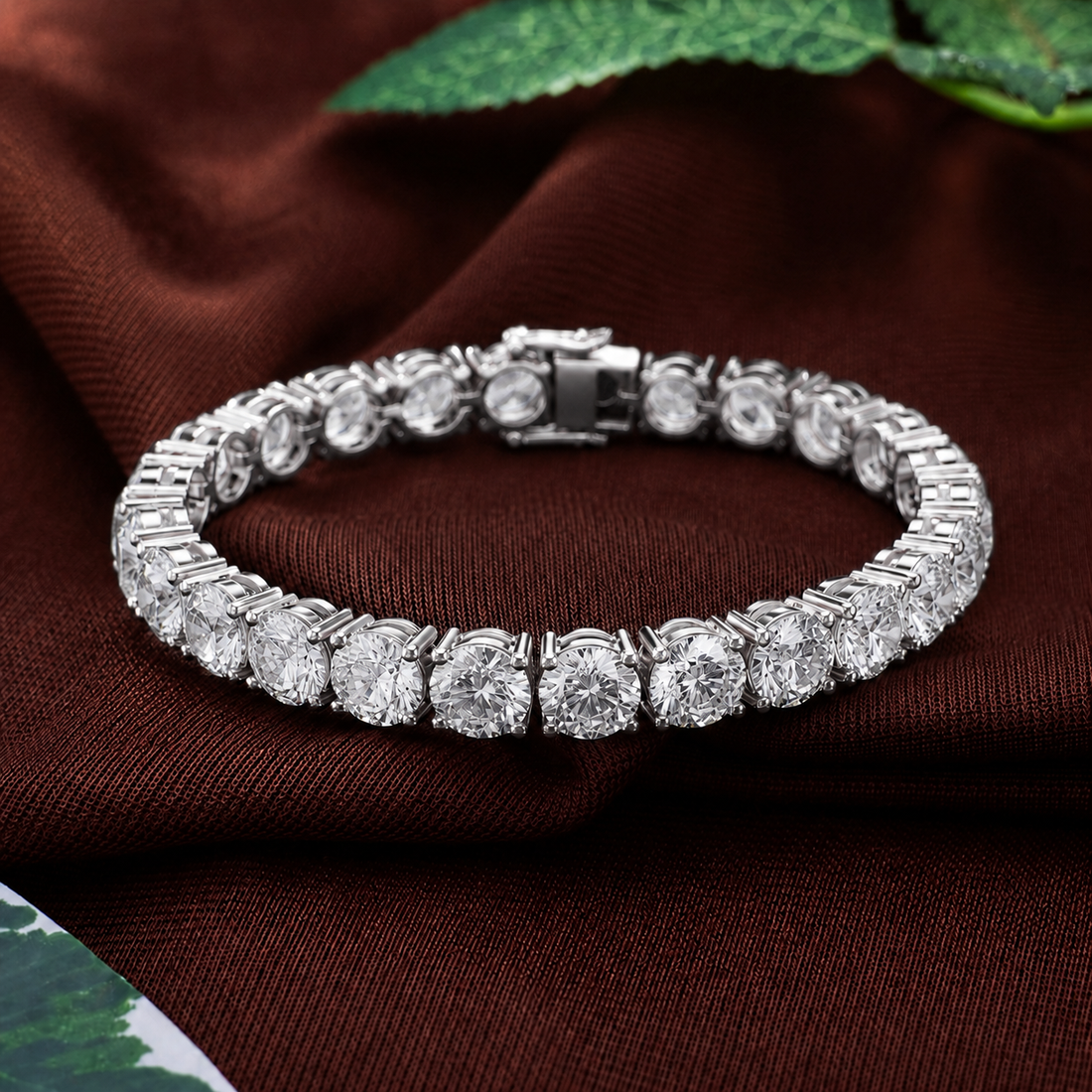 A silver-toned tennis bracelet with round-cut clear stones, displayed on a draped dark brown fabric background with a small green leaf in the corner.