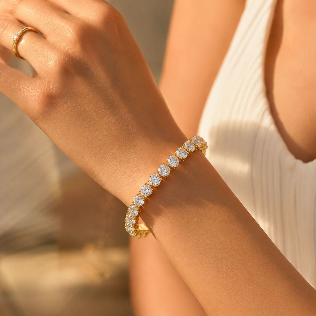 A gold tennis bracelet with round clear stones worn on a woman's wrist, shown in warm sunlight against a soft neutral background.
