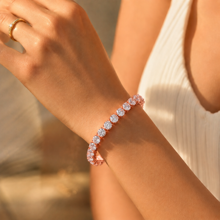A close-up lifestyle shot of a woman's wrist wearing a rose gold tennis bracelet with sparkling clear gemstones. The warm sunlight highlights the jewelry, and a matching rose gold ring is visible on her finger.