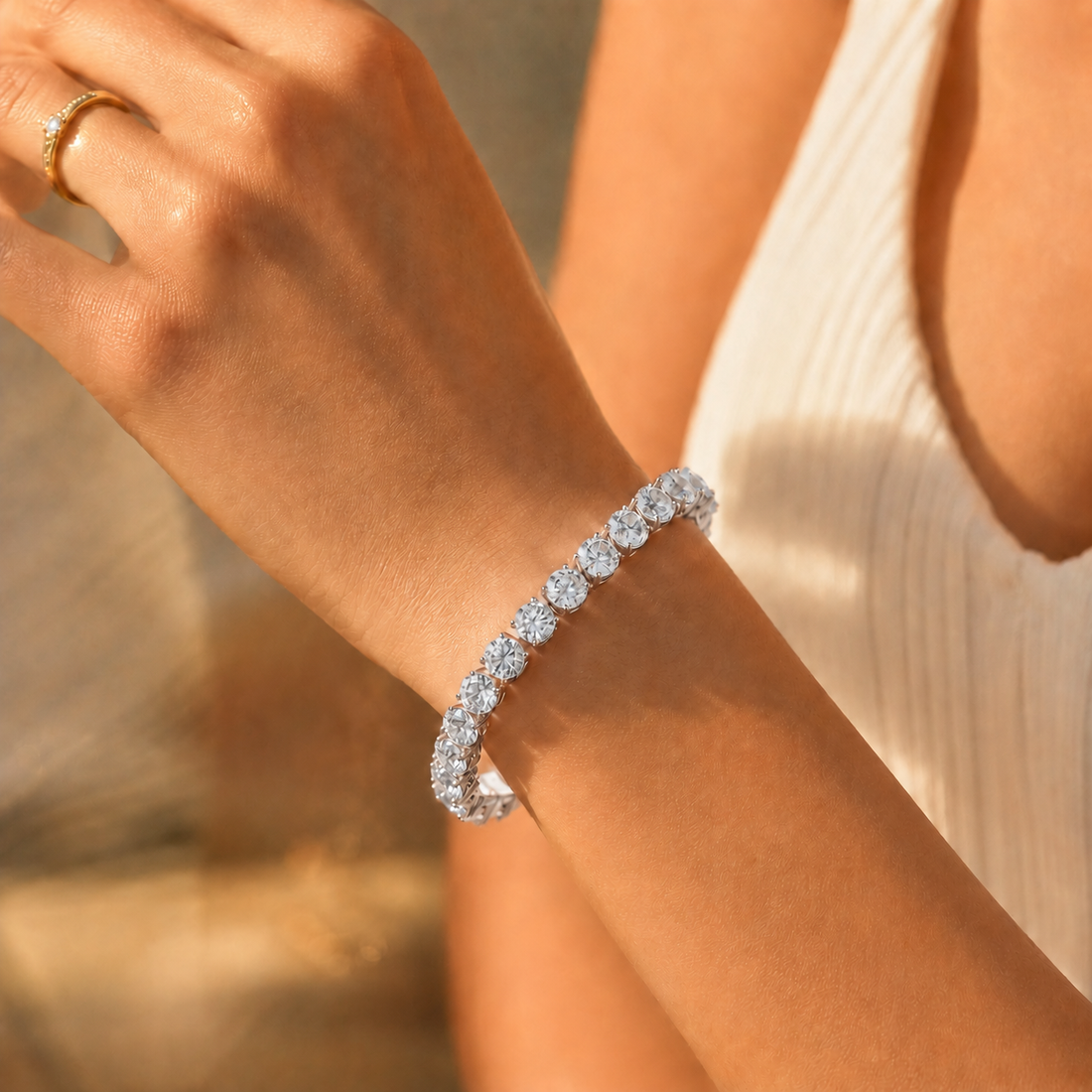 A close-up of a woman's wrist wearing a sparkling diamond tennis bracelet, with a gold ring on her finger and a white ribbed top in the background.