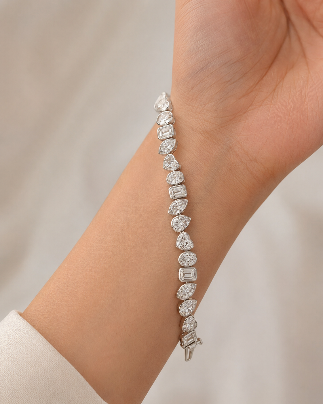 A close-up of a diamond tennis bracelet on a person's wrist, featuring a silver-toned clasp and multiple diamond-cut stones in pear, marquise, emerald, and round shapes.