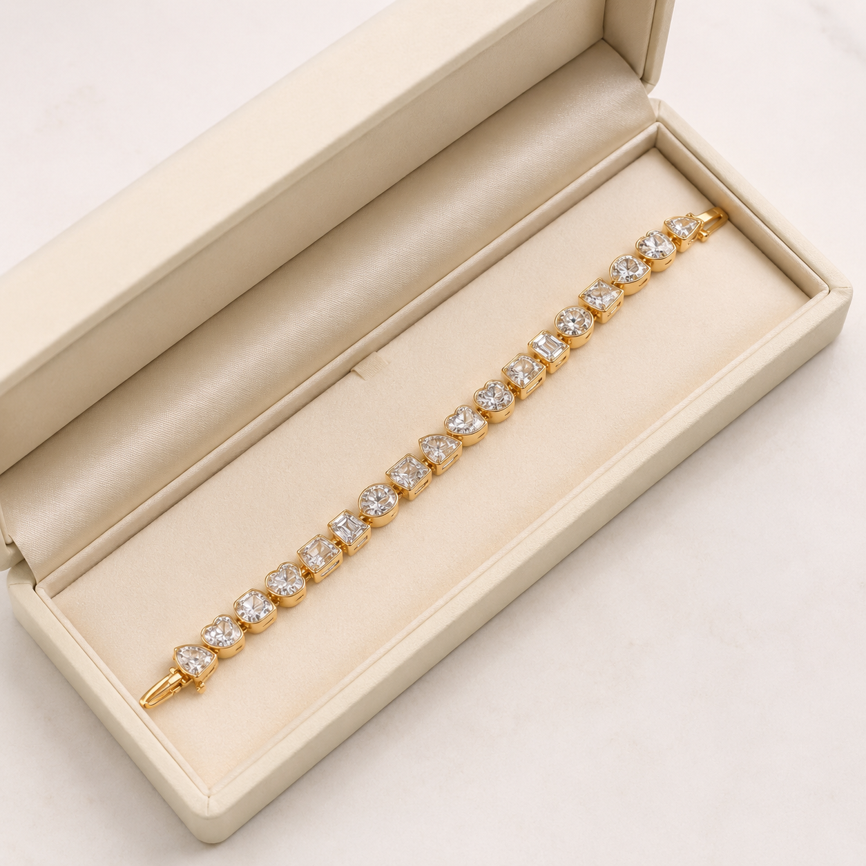 A gold bracelet with round clear gemstones displayed inside an open cream-colored jewelry box on a light marble surface.