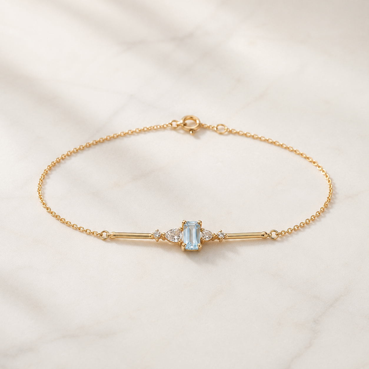 A delicate gold chain bracelet with a central bar design, featuring a light blue rectangular gemstone and small clear accent stones, displayed on a light marble surface.