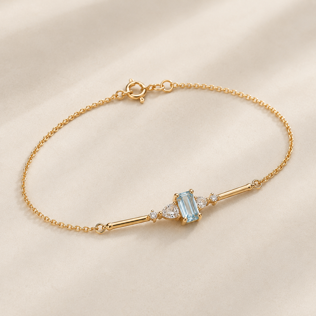 A delicate gold chain bracelet featuring a central light blue emerald-cut gemstone flanked by small clear stones, displayed on a soft beige fabric background.