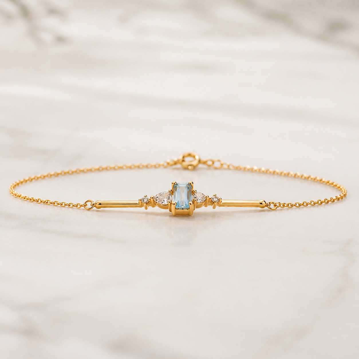 A delicate gold chain bracelet with a central bar design, featuring a light blue rectangular gemstone and small clear accent stones, displayed on a light marble surface.