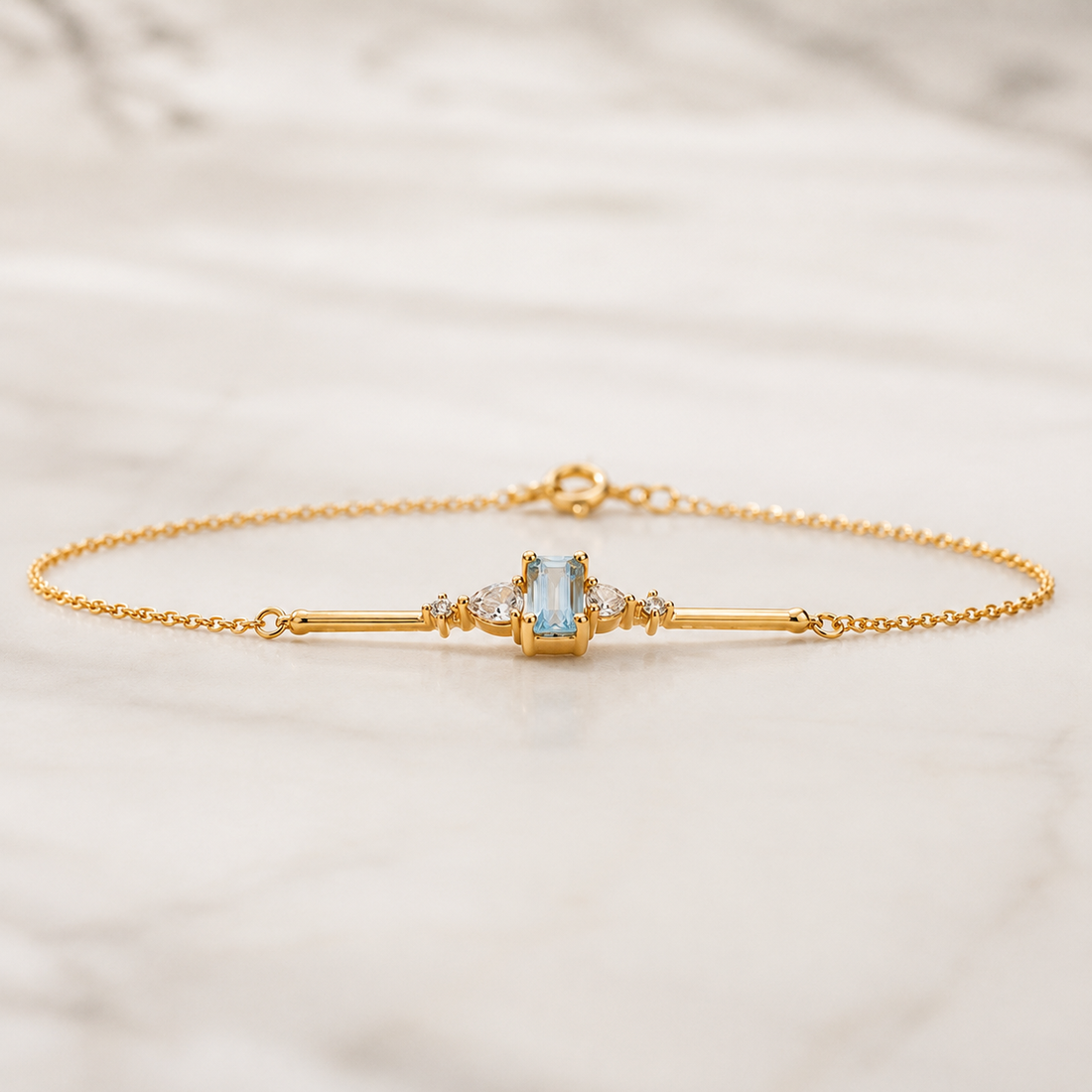 A delicate gold chain bracelet with a central bar design, featuring a light blue rectangular gemstone and small clear accent stones, displayed on a light marble surface.