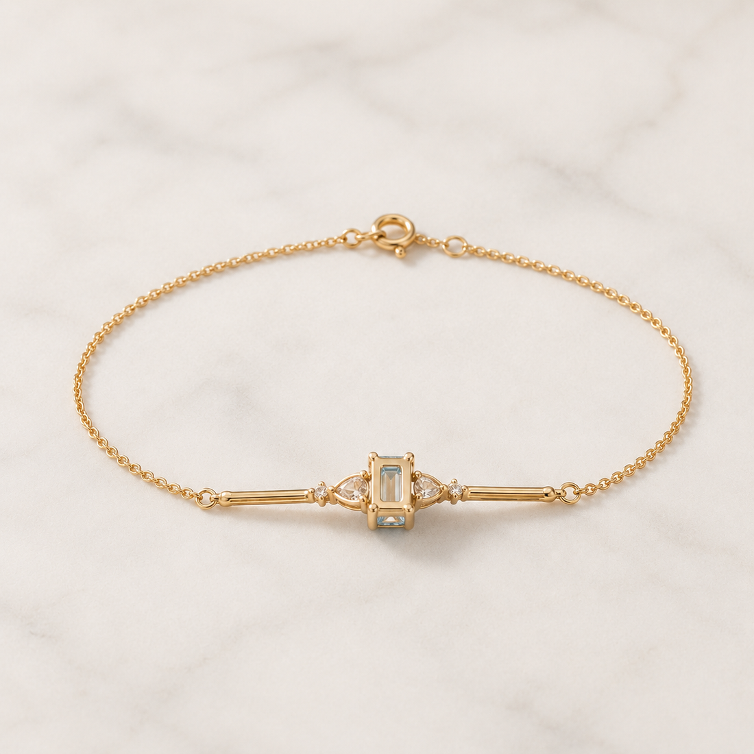 A delicate yellow gold chain bracelet with a central baroque-style bezel-set oval light blue stone, accented by small clear stones, displayed on a white marble surface.
