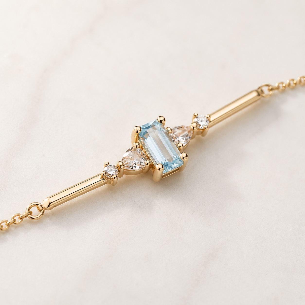 A gold bar bracelet featuring a central light blue baguette-cut gemstone, flanked by small clear round and heart-shaped stones, displayed on a light marble surface.