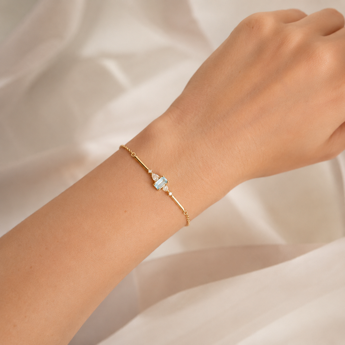 A delicate gold chain bracelet with a central light blue rectangular stone and small clear accent stones, shown worn on a person's wrist against a soft white fabric background.