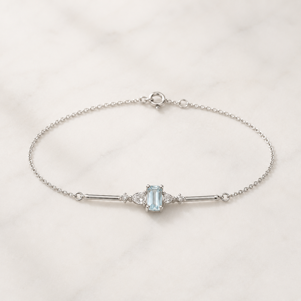 A delicate silver chain bracelet featuring a central light blue emerald-cut gemstone flanked by small clear stones, displayed on a white marble surface.