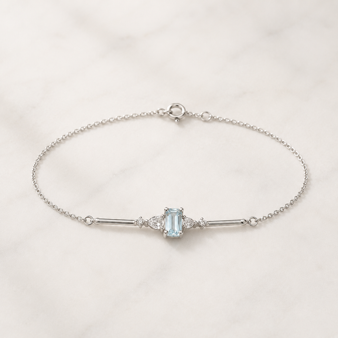 A delicate silver chain bracelet featuring a central light blue emerald-cut gemstone flanked by small clear stones, displayed on a white marble surface.