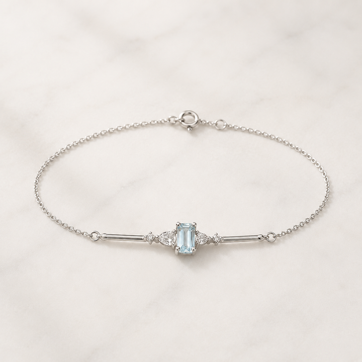 A delicate silver chain bracelet featuring a central light blue emerald-cut gemstone flanked by small clear stones, displayed on a white marble surface.