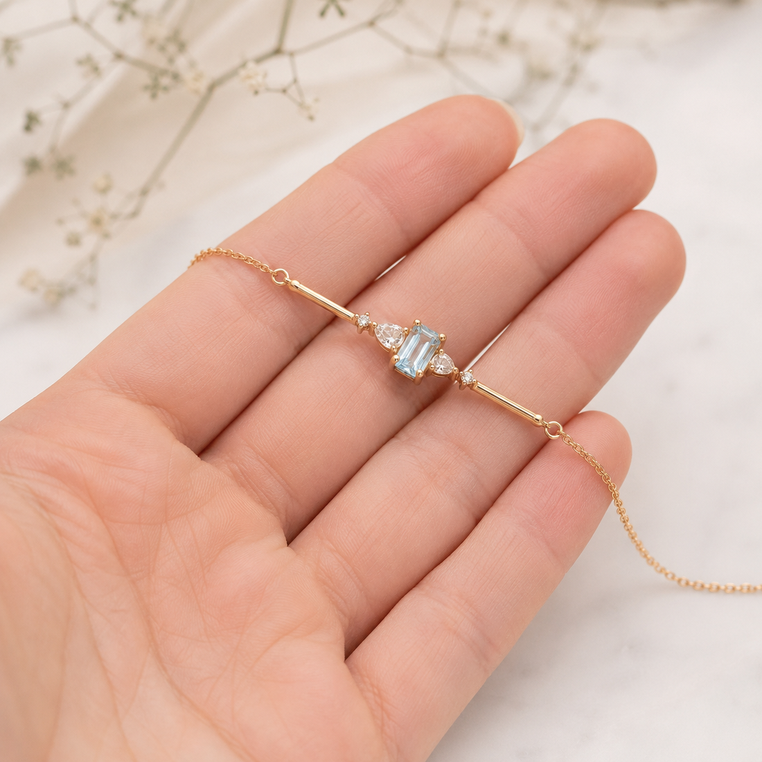 A delicate gold chain bracelet with a central light blue rectangular gemstone and small clear accent stones, shown resting on a person's hand against a soft white background with faint dried flowers.