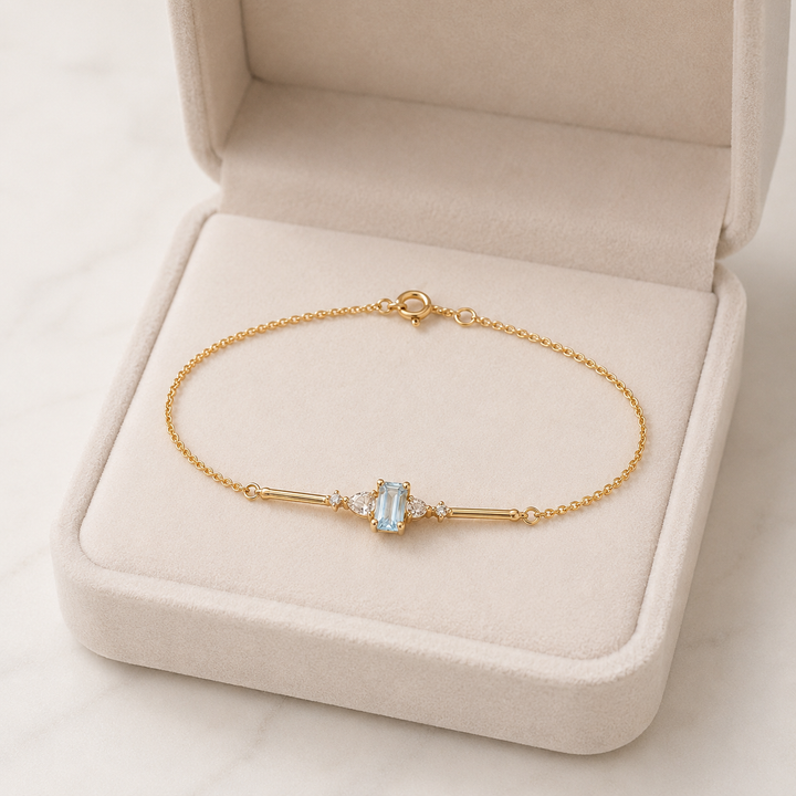 A delicate gold chain bracelet with a central light blue rectangular gemstone and small clear stones, displayed on a beige jewelry box.
